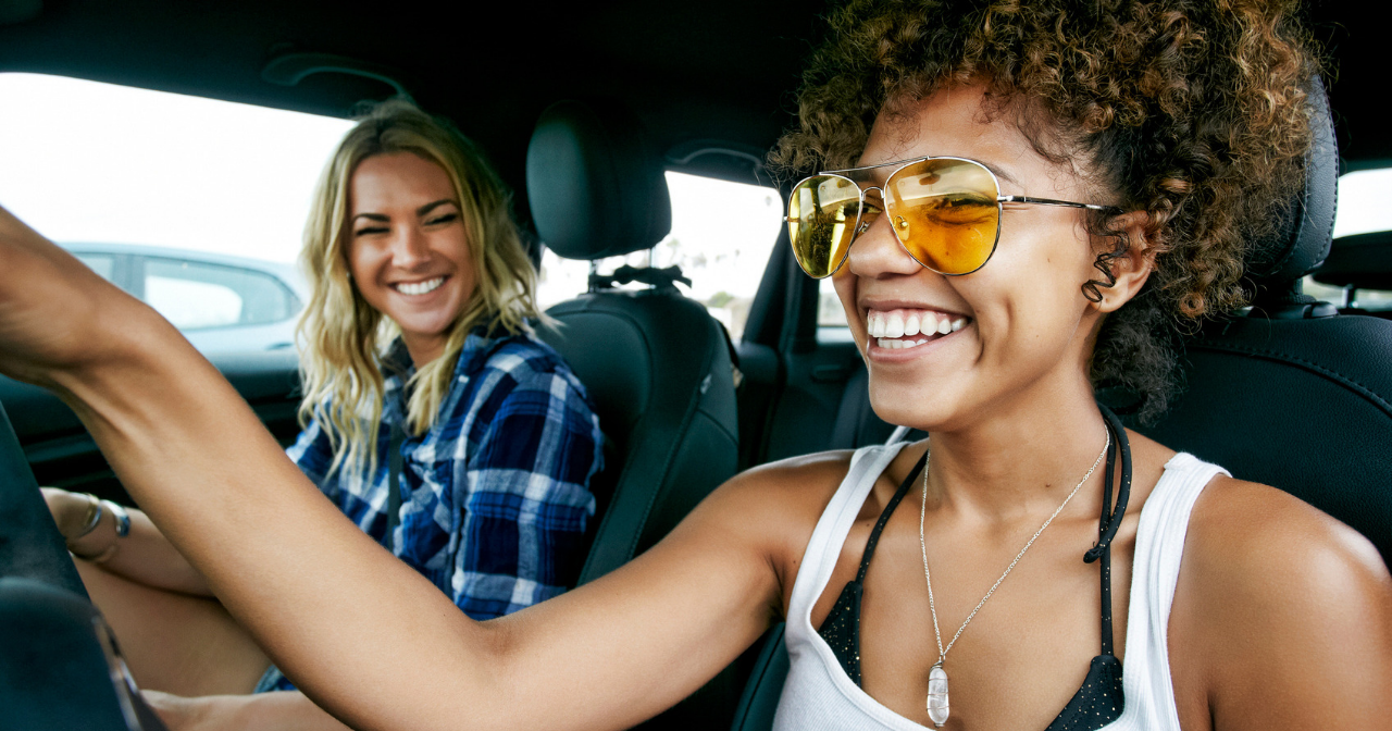 Portrait of two women with long blond and brown curly hair sitting in car, wearing sunglasses, smiling.