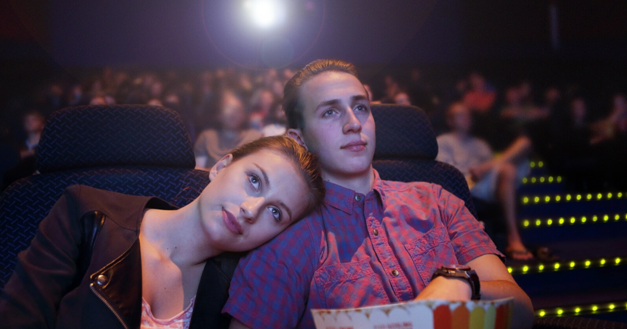 Two teens snuggle while watching a movie at the theatre. 