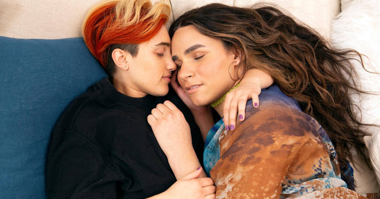 A gender nonbinary/trans couple lay in bed together and snuggle. 