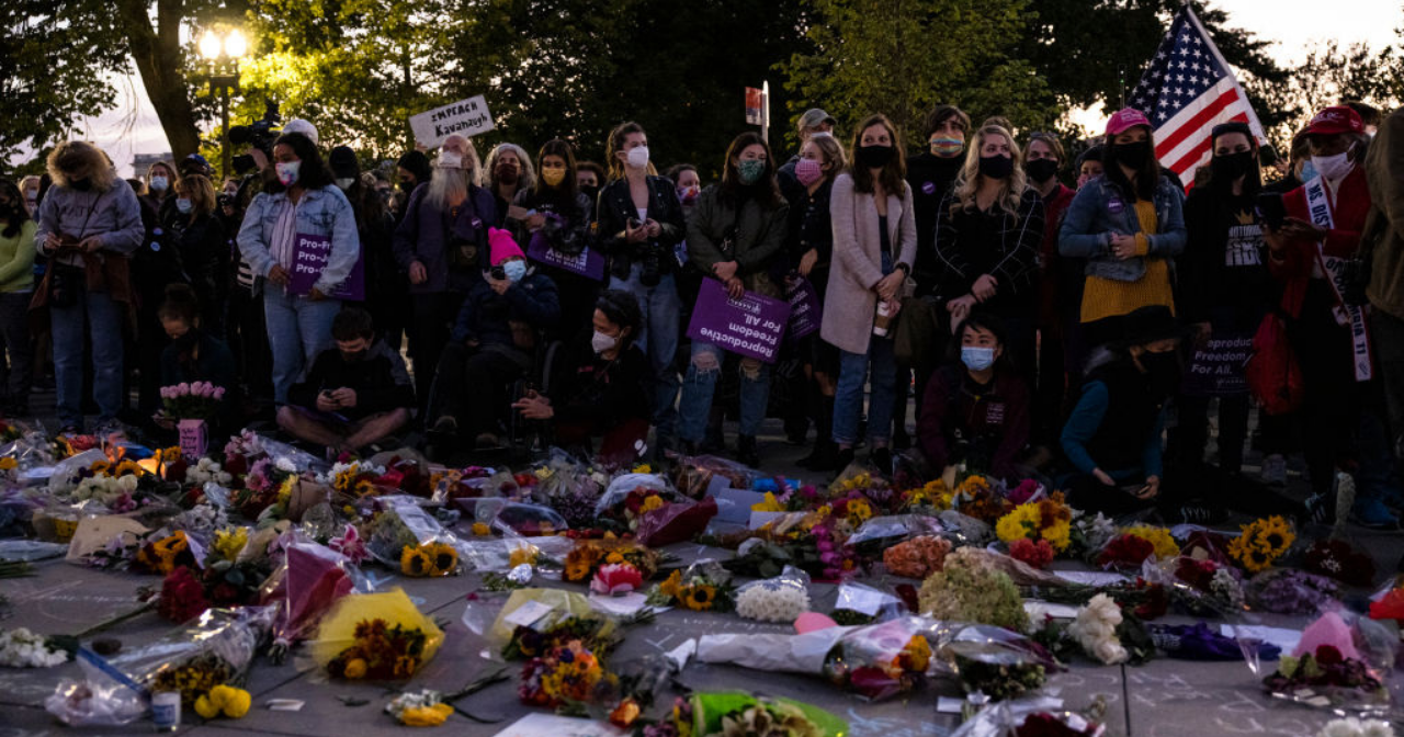 A group of mostly women standing in front of flowers and posters outside the Supreme Court. 