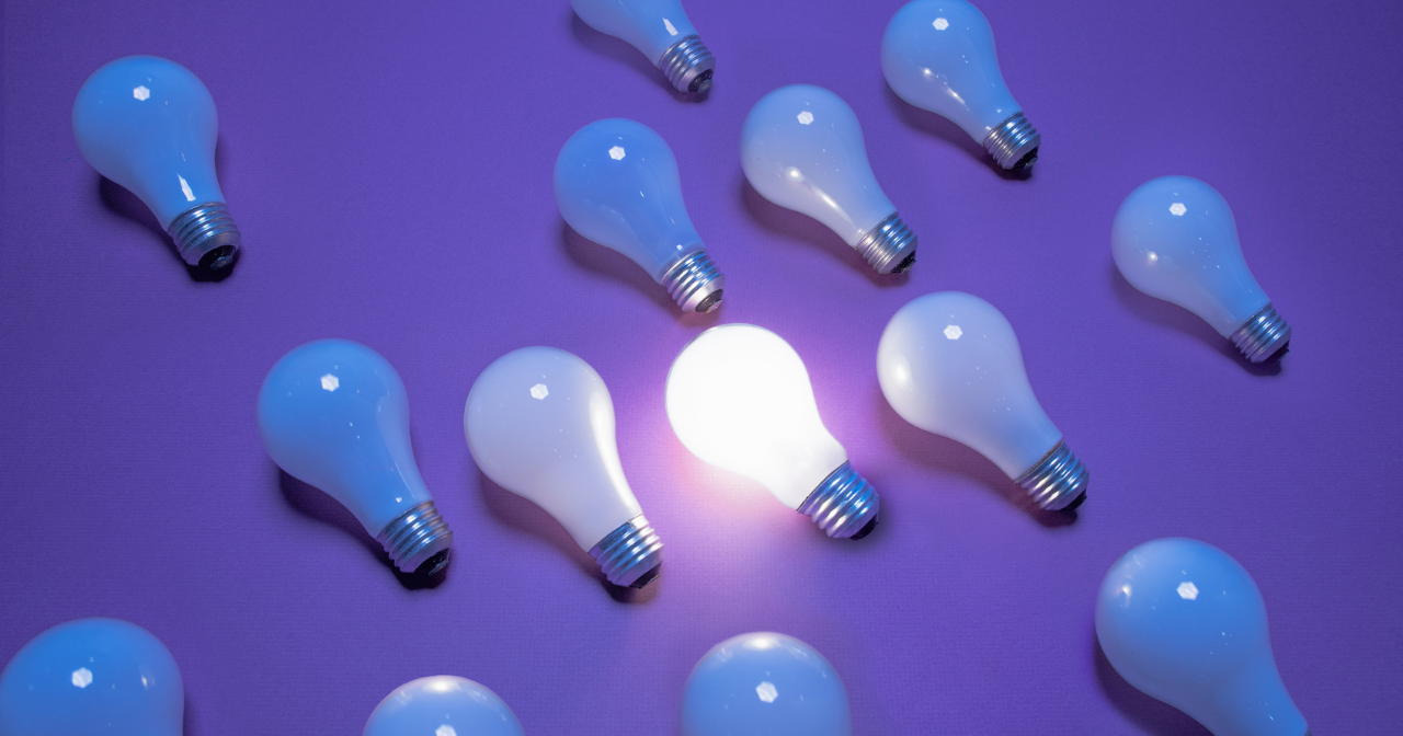 A group of lightbulbs lay on a purple background with only one lit up. 