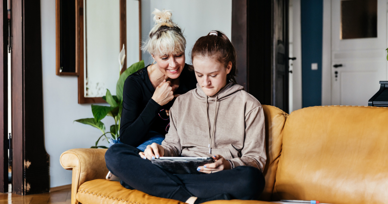 A mother and her teenage daughter sit on the sofa with an ipad. 