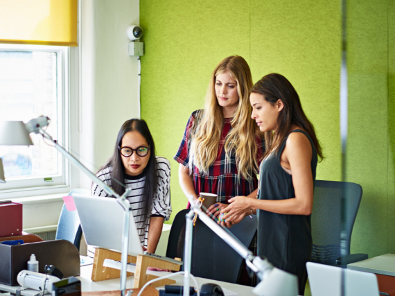 Three women gather around a computer in an office