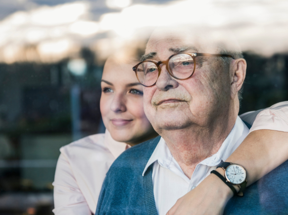A woman hugs her grandfather from behind as they both look out a window. 