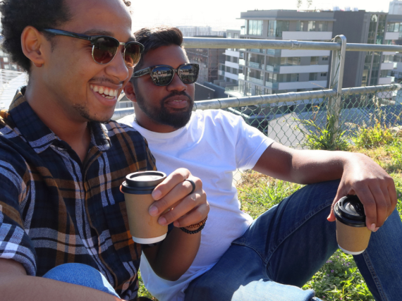 Two Black men relax on a rooftop with coffee. 
