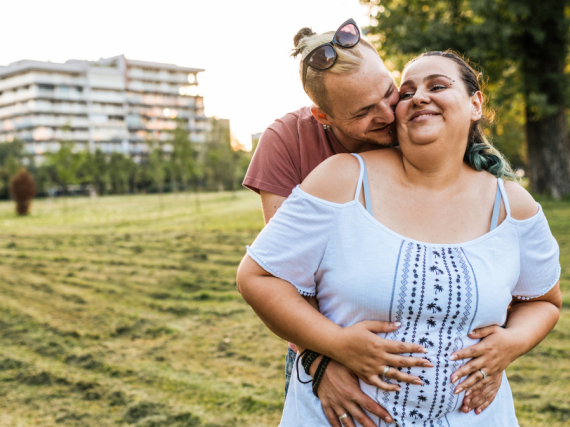 A pregnant woman smiles and is happily hugged by her male partner. 