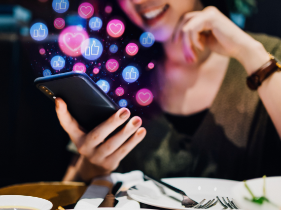 A woman looks at her phone while eating a meal and images of social media icons float out from the screen. 
