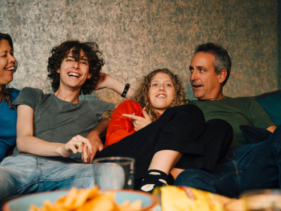A family of four laugh on the sofa while watching TV. 