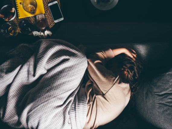 A woman lays curled up under a blanket on a sofa while holding her head and overwhelmed. 